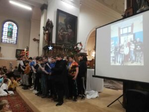 Dens enfants célèbrent Noël dans une église en chantant. Sur un grand écran, une photo de Saint-Nicolas dans une classe